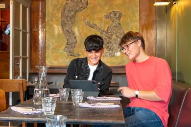 Zwei junge Männer, die ein JRK-Tandem bilden, sitzen an einem Tisch in einem Café und schauen gemeinsam auf ein iPad.