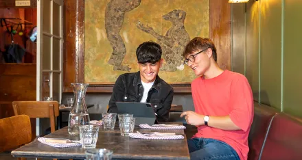 Zwei junge Männer, die ein JRK-Tandem bilden, sitzen an einem Tisch in einem Café und schauen gemeinsam auf ein iPad.