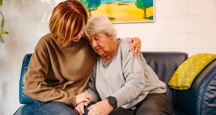 Eine ältere Frau und ihre Tochter sitzen auf dem Sofa. Die jüngere Frau umarmt ihre Mutter.