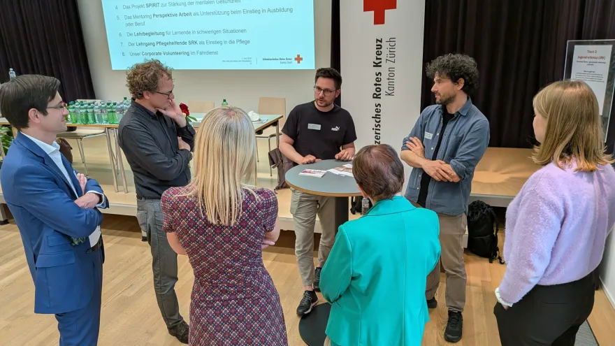 Rund um einen Stehtisch stehen mehrere Personen und hören gespannt zu. Es wird über ein Programm vom Jugendrotkreuz Zürich erzählt. 