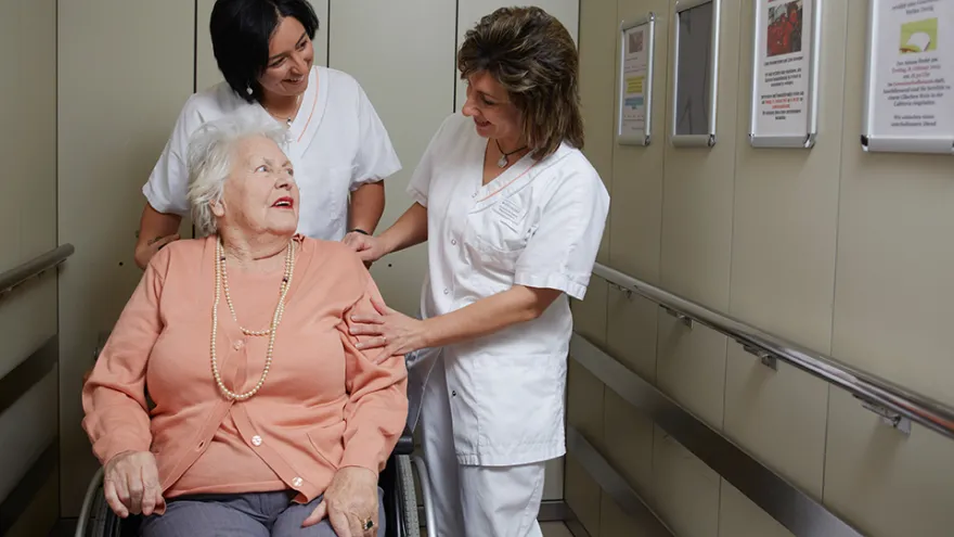 Zwei Pflegehelferinnen kümmern sich um eine Patientin im Rollstuhl