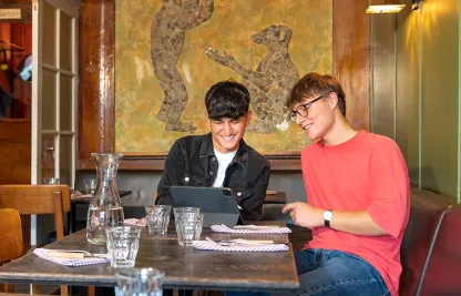 Zwei junge Männer, die ein JRK-Tandem bilden, sitzen an einem Tisch in einem Café und schauen gemeinsam auf ein iPad.